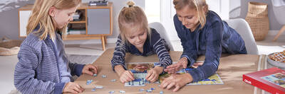 Drei Kinder puzzlen gemeinsam am Tisch an einem großen Motiv, auf dem sie selbst abgebildet sind.