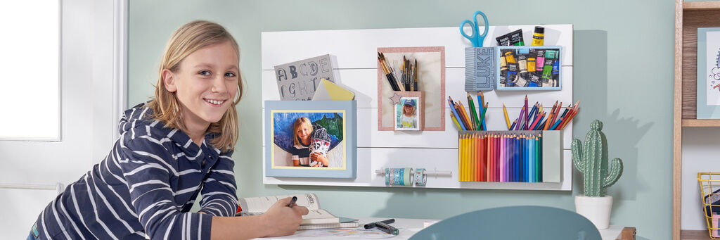 Kind steht neben Schreibtisch. Darüber hängt ein Brett mit verschiedenen kleinen befestigten Behältern, in denen sich Schreib- und Bastelutensilien befinden.