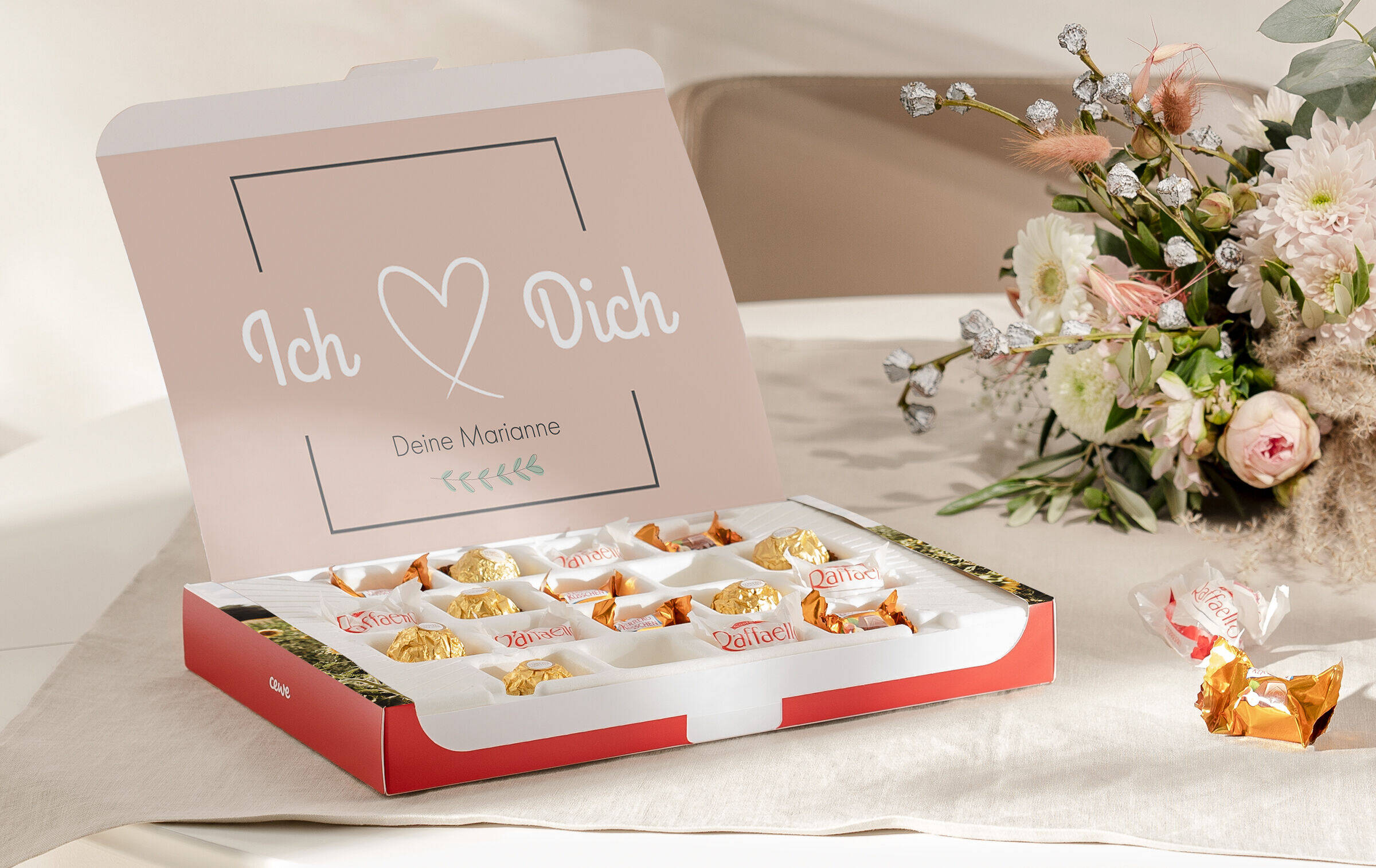 Die aufgeklappe Foto-Geschenkbox steht neben einem Blumenstrauß auf einem Tisch. Auf der Innenseite der Box steht mit Cliparts und Text "Ich liebe Dich. Deine Marianne".