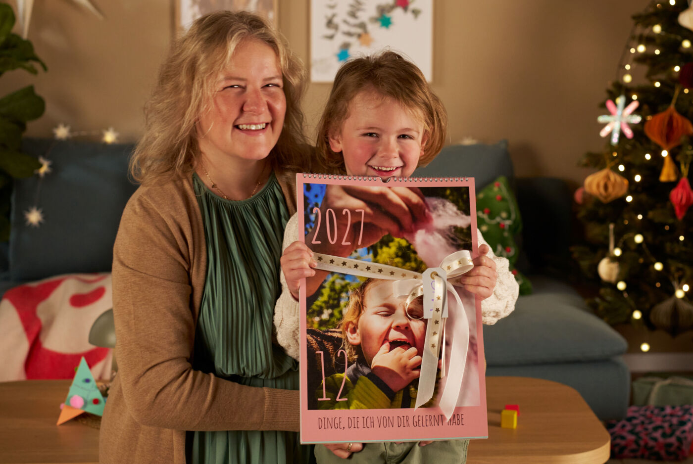 Ein Kind uns seine Mama stehen in einem Wohnzimmer neben einem geschmückten Weihnachtsbaum. Der Junge hält einen Fotokalender in die Kamera.
