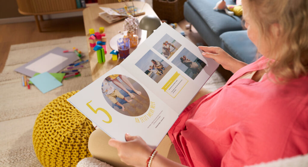Eine Frau sitzt auf einem Sofa und blättert durch ein CEWE FOTOBUCH, die Doppelseite zeigt Fotomotive und die Zahl „5“.
