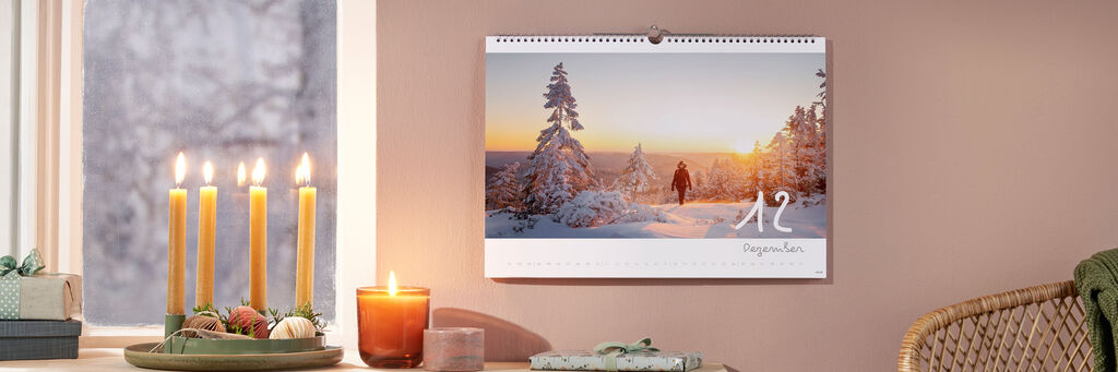 An der Wand hängt ein Kalender von Annika und Mathias Koch. Zu sehen ist der Monat Dezember, der eine Frau (Annika) zeigt, die bei Sonnenuntergang durch eine Winterlandschaft läuft. Rechts unterhalb des Kalenders steht ein Korbstuhl mit einer grünen Decke. Links ist unter dem Fenster ein Sideboard erkennbar, auf dem Adventskerzen und ein Geschenk zu sehen sind.