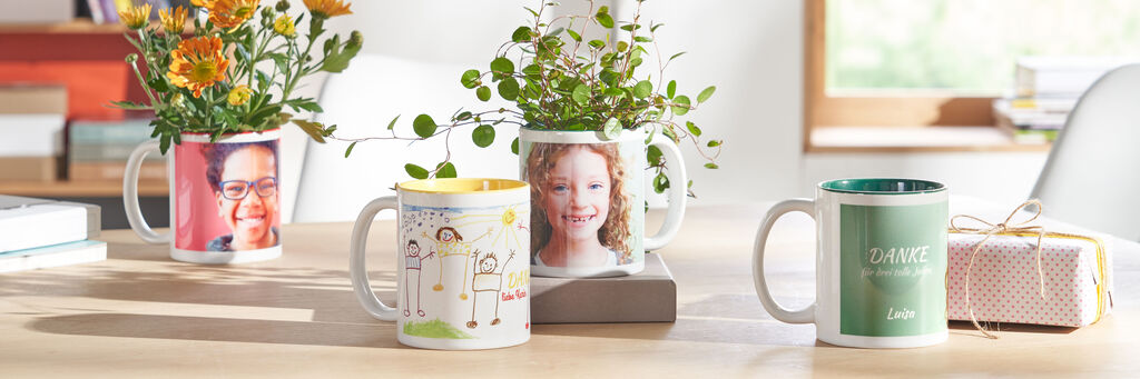 Auf einem Tisch stehen mehrere Fototassen. Darauf sind Fotos von Kindern und selbst gemalte Kinderbilder zu sehen. In zwei der Fototassen befinden sich Grünpflanzen, daneben liegt ein Geschenk.