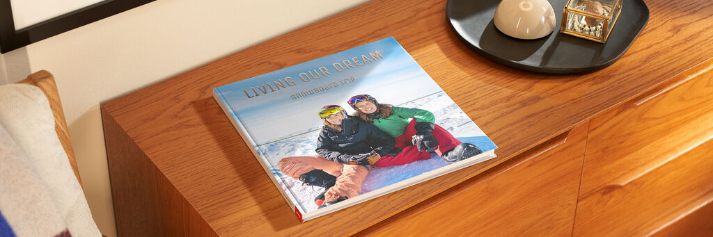 Auf einem Holzschrank liegt ein geschlossenes CEWE FOTOBUCH. Auf dem Cover sind zwei Frauen in Ski-Anzügen zu sehen, die in einer verschneiten Landschaft sitzen. Die Überschrift lautet „Living our dream – Snowboard Trip“.
