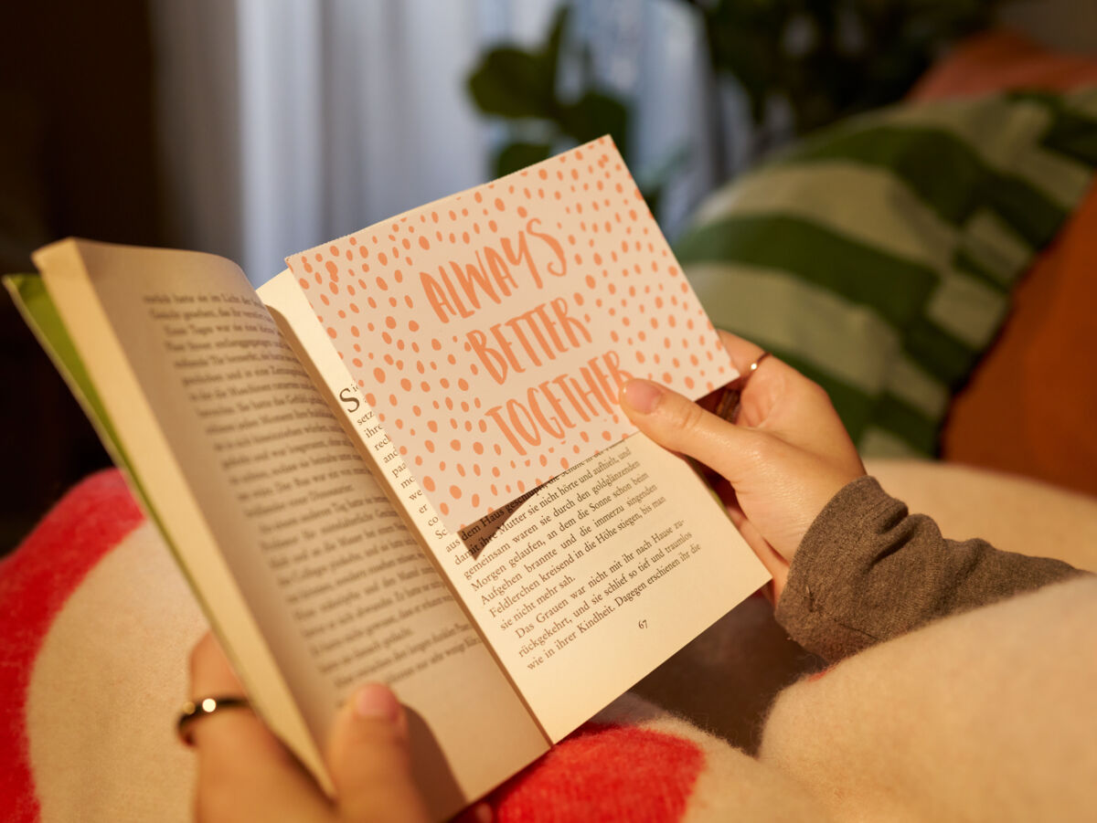 Eine Frau sitzt auf einem Sofa und hält ein aufgeklapptes Buch in den Händen, zwischen den Seiten hält sie eine Fotopostkarte.