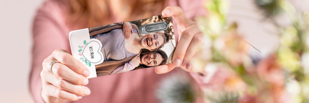 Eine Frau fotografiert mit ihrem Handy Richtung Betrachter. Geschützt wird das Handy mit einer Hülle. Auf diesem sind sind Mutter und Tochter zu sehen, die gemeinsam in die Kamera lachen.