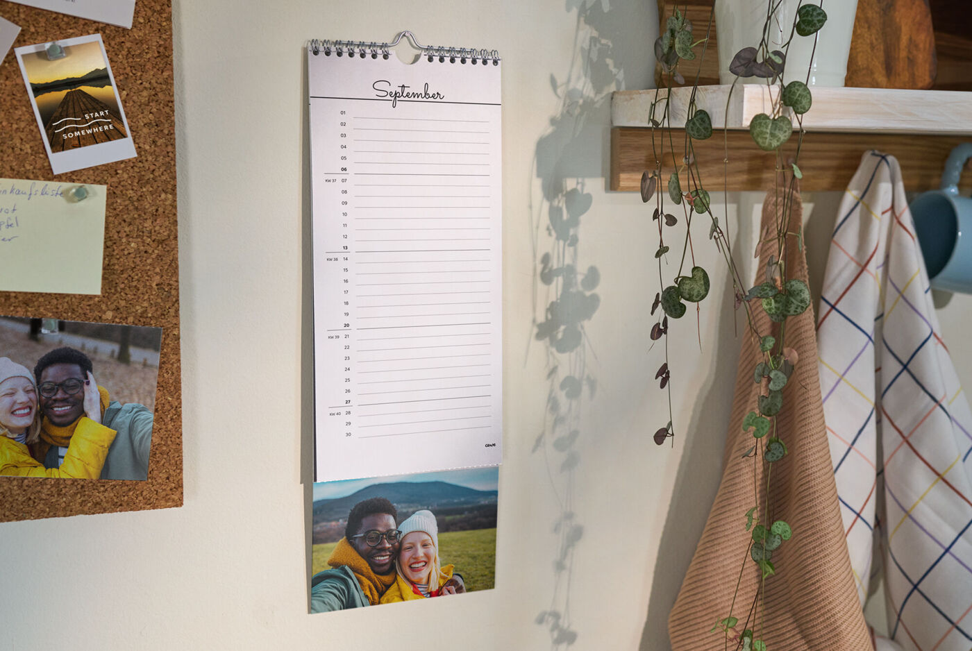 Ein Küchenkalender mit abtrennbaren Foto am Ende hängt an einer weißen Küchenwand neben einem Regal und einer Pinnwand.